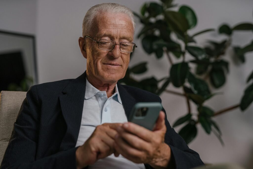Elderly man in glasses smiles while using a smartphone at home.